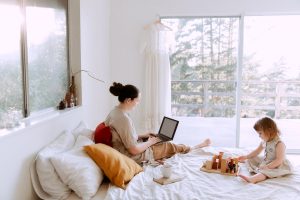 adorable toddler girl playing with wooden blocks sitting on bed while mother using laptop on sunny morning